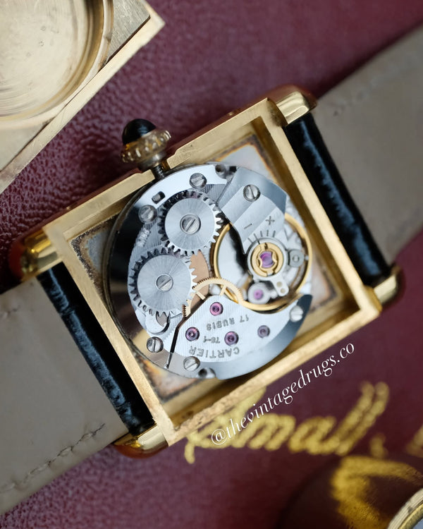Close-up of a Cartier watch showing the open back and exposed mechanical movement, with a gold-tone case, silver gears, and a beige leather strap on a dark burgundy textured surface.